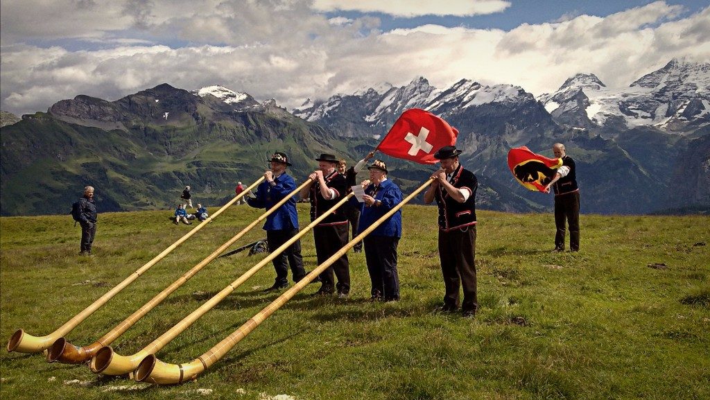 Alphorn players in Swiss dress performing on an alpine meadow below snow-covered peaks, with the Swiss flag flying